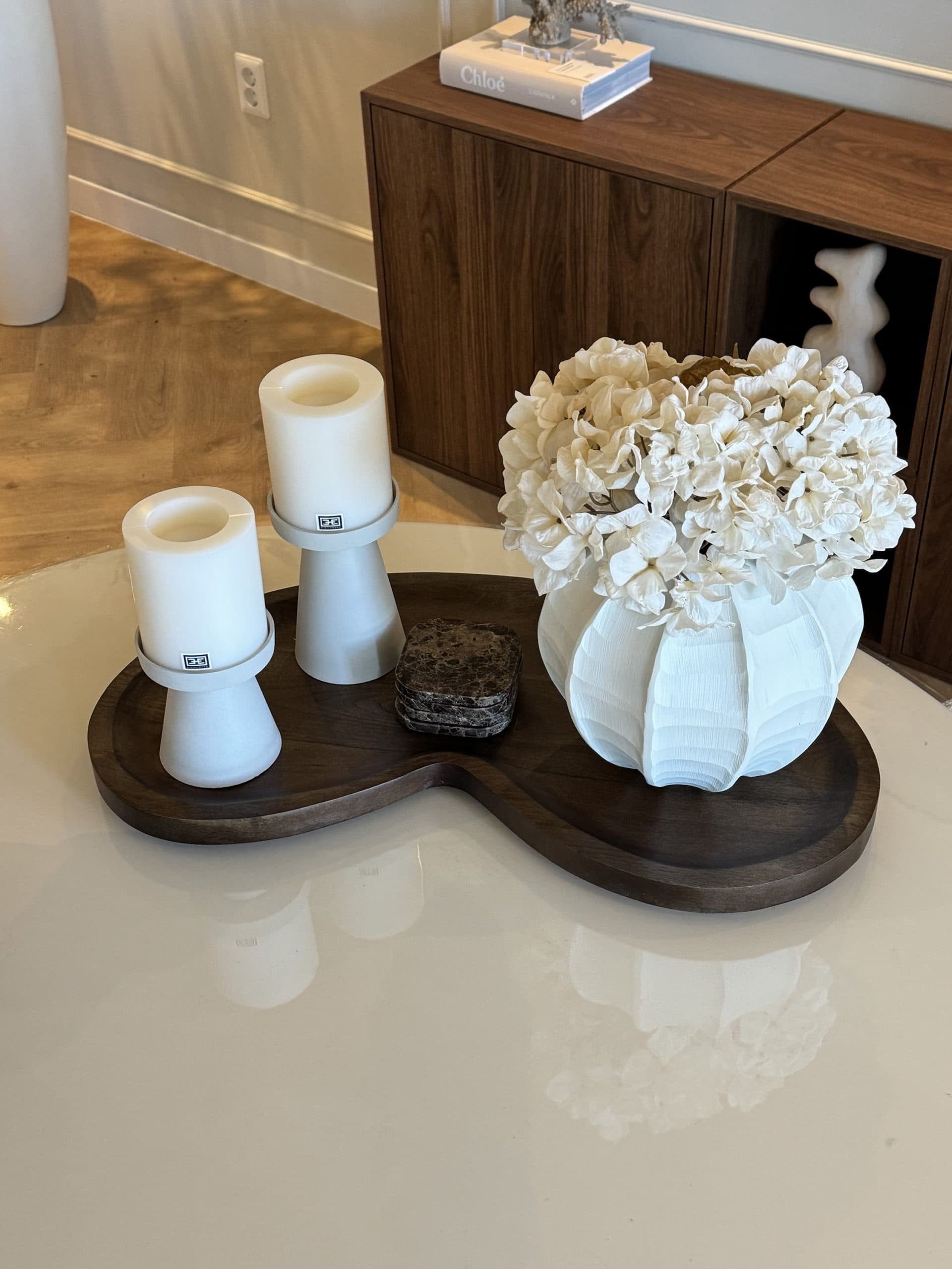 Donkerhouten dienblad op een salontafel met twee witte kaarshouders, marmeren onderzetters en een witte ribbelvaas met hortensia's